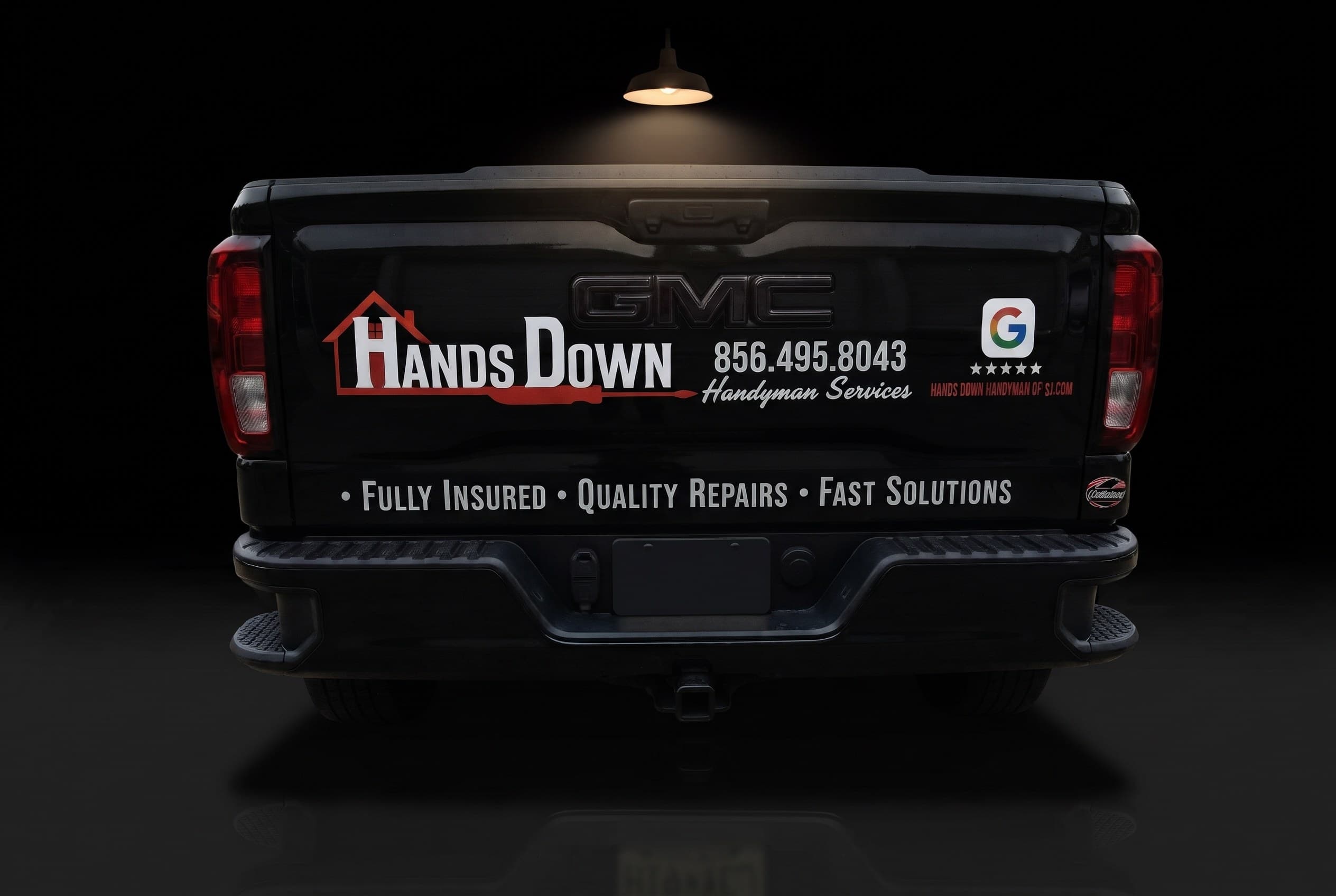 The tailgate of Hands Down Handyman's black GMC pickup under a single barn light — the logo, (856) 495-8043 and the Google five-star badge lettered across it